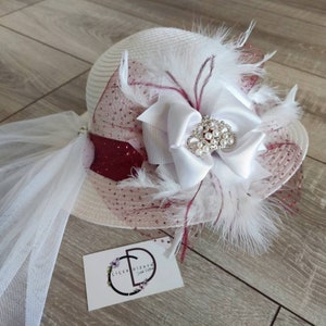 May include: White hat with a white bow and a pearl crown, decorated with white feathers and a burgundy tulle veil. The hat is sitting on a wooden surface with a business card that says "Cicek Dizayn" in white text.