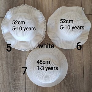 May include: Three white straw sun hats with different sizes. The hats are labeled with their size in centimeters and the age range they are suitable for. The hats are on a wooden floor.