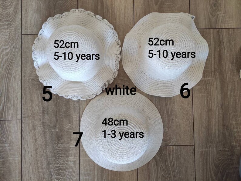 May include: Three white straw sun hats with different sizes. The hats are labeled with their size in centimeters and the age range they are suitable for. The hats are on a wooden floor.