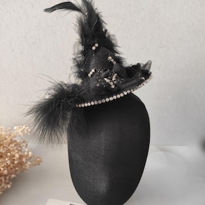 May include: A black witch hat with a decorative design. The hat features a brim lined with small, sparkling rhinestones and is adorned with black feathers and tulle. The hat is displayed on a black mannequin head.