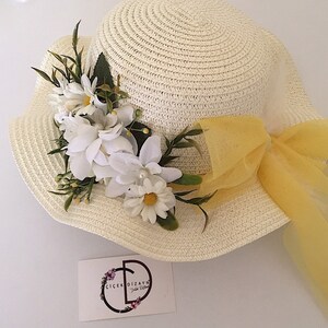 May include: A white straw hat with a yellow ribbon and a white flower embellishment. The hat has a wide brim and a small crown. The flower embellishment is made of white fabric flowers and green leaves.