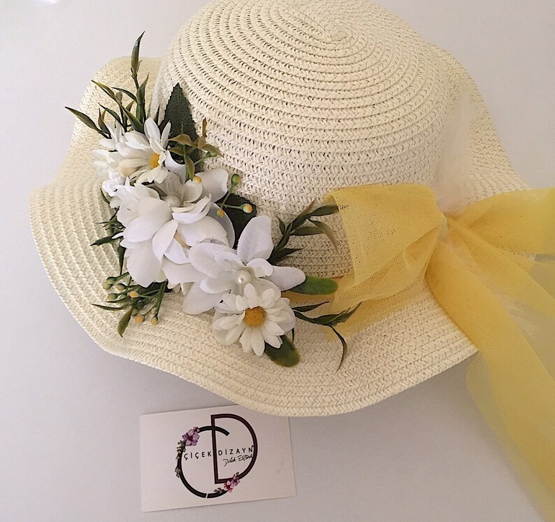 May include: A white straw hat with a yellow ribbon and a white flower embellishment. The hat has a wide brim and a small crown. The flower embellishment is made of white fabric flowers and green leaves.