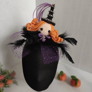 May include: A handmade Halloween ornament featuring a crocheted witch with orange hair and a black hat adorned with rhinestones. The witch is set on a black fabric base with purple accents and black feathers. Two small orange pumpkins are in the background.