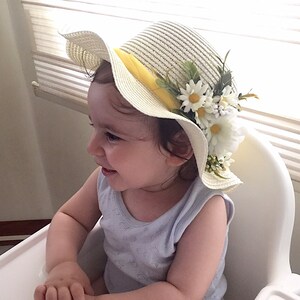 May include: A young child wearing a white straw hat with a yellow ribbon and white flowers. The hat has a wide brim and is decorated with green leaves.