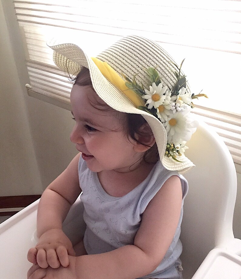 May include: A young child wearing a white straw hat with a yellow ribbon and white flowers. The hat has a wide brim and is decorated with green leaves.