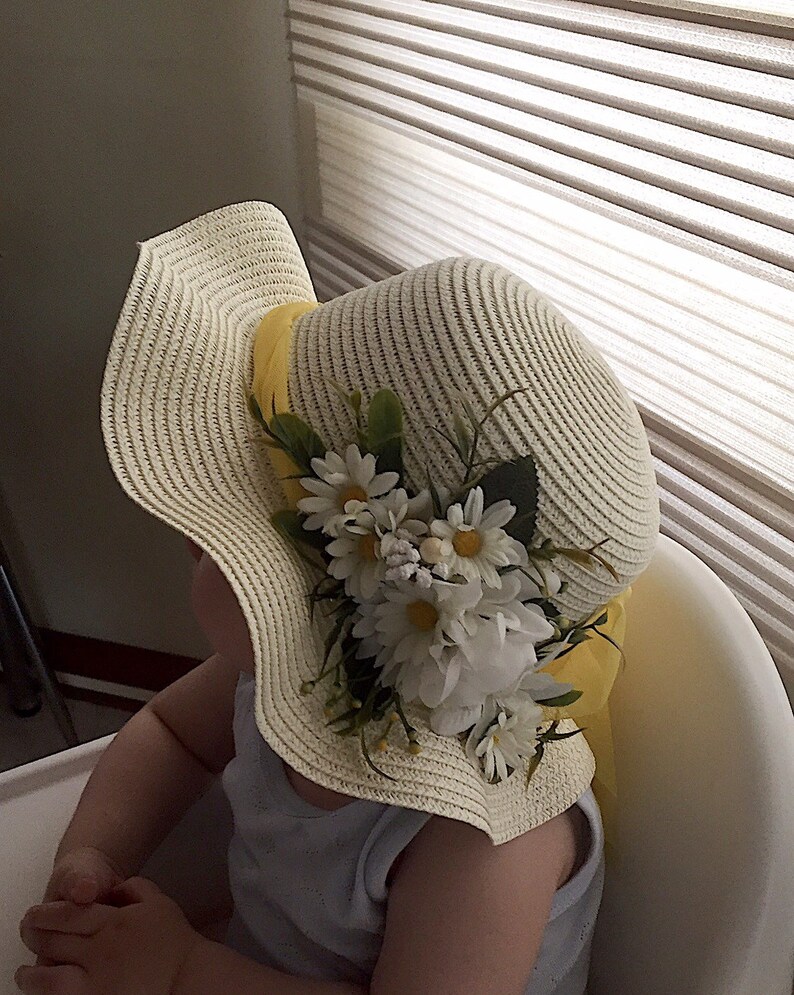 May include: A white straw sun hat with a yellow ribbon and white flowers. The hat is worn by a child sitting in a chair.