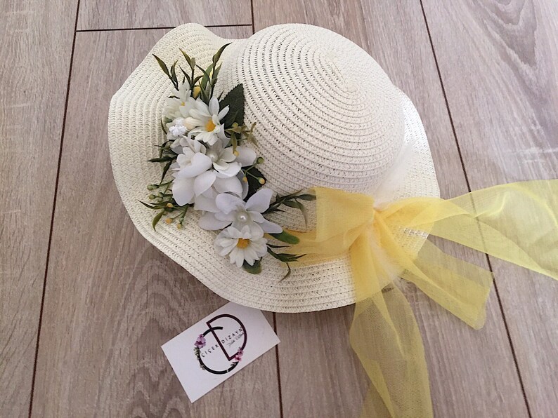 May include: A white straw hat with a yellow ribbon and white flowers. The hat is on a wooden surface.