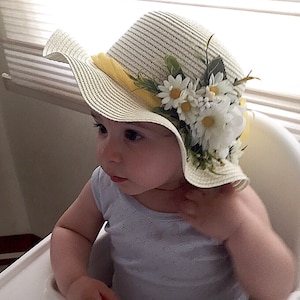 May include: A baby wearing a white straw hat with a yellow ribbon and white daisy flowers. The hat has a wide brim and is decorated with green leaves.