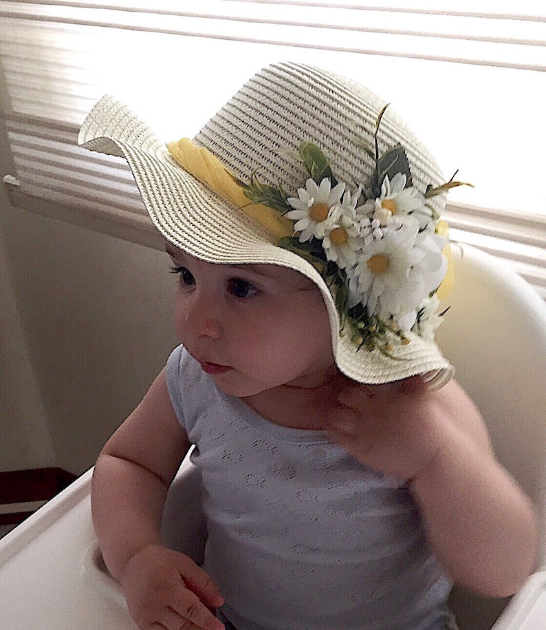 May include: A baby wearing a white straw hat with a yellow ribbon and white daisy flowers. The hat has a wide brim and is decorated with green leaves.