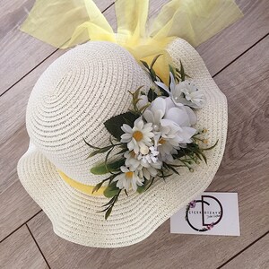 May include: A white straw hat with a yellow ribbon and a white flower embellishment. The hat is decorated with a yellow tulle bow. The hat is sitting on a wooden surface.  The text on the card reads "&Ccedil;i&ccedil;ek Dizayn"