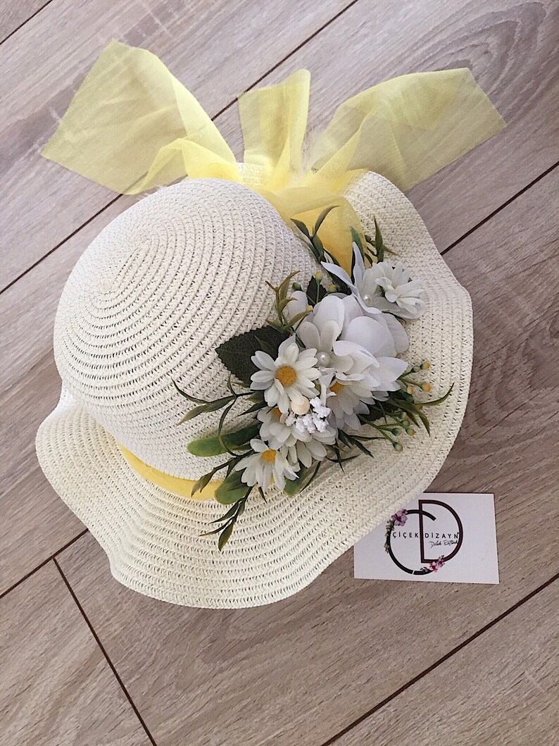 May include: A white straw hat with a yellow ribbon and a white flower embellishment. The hat is decorated with a yellow tulle bow. The hat is sitting on a wooden surface.  The text on the card reads "&Ccedil;i&ccedil;ek Dizayn"