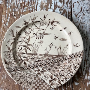 May include: A round ceramic plate featuring a brown transferware pattern. The design depicts a landscape with bamboo, birds, and water lilies, while the lower section displays a geometric and floral patchwork border in brown and cream.