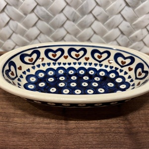 May include: A white oval ceramic dish with a blue and brown heart pattern. The dish has a white rim with a blue and white polka dot pattern.