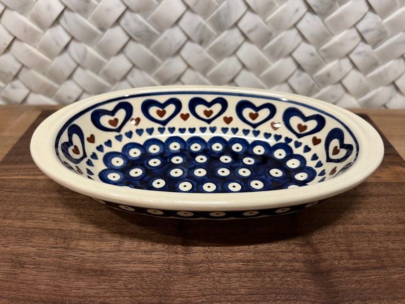 May include: A white oval ceramic dish with a blue and brown heart pattern. The dish has a white rim with a blue and white polka dot pattern.