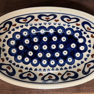 May include: An oval ceramic serving dish with a white background and blue and brown designs. The dish features a pattern of blue circles with white centers, surrounded by a border of brown hearts and blue triangles.