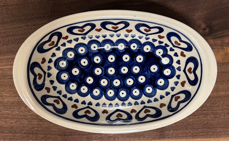 May include: An oval ceramic serving dish with a white background and blue and brown designs. The dish features a pattern of blue circles with white centers, surrounded by a border of brown hearts and blue triangles.