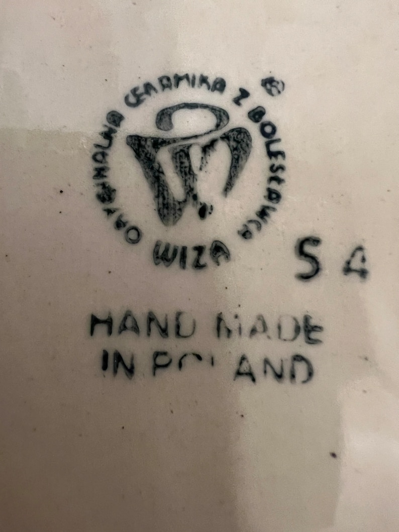 May include: A white ceramic plate with a black stamp that reads "Ceramika z Boleslawca" and "Wiza" with a stylized "W" logo. The plate also has the text "HAND MADE IN POLAND" and the number "54" printed on it.