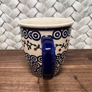 May include: A blue and white ceramic mug with a blue handle. The mug features a pattern of circles and brown berries on a vine.