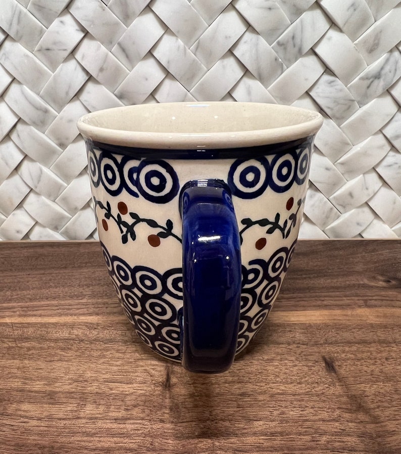 May include: A blue and white ceramic mug with a blue handle. The mug features a pattern of circles and brown berries on a vine.