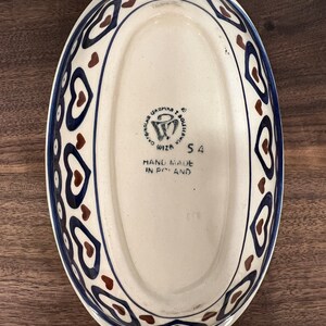 May include: White oval ceramic baking dish with a blue and brown heart pattern around the rim. The bottom of the dish has a brown stamp that reads "Ceramika Boleslawiec,  Wiza,  Hand Made in Poland,  54".