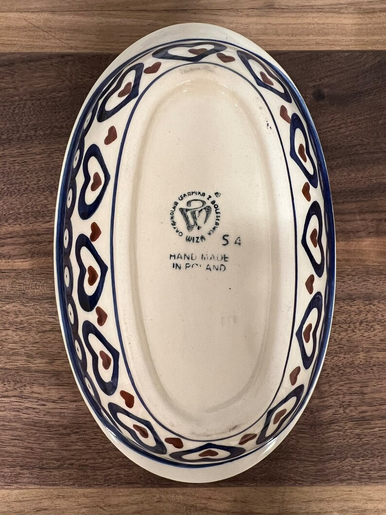 May include: White oval ceramic baking dish with a blue and brown heart pattern around the rim. The bottom of the dish has a brown stamp that reads "Ceramika Boleslawiec,  Wiza,  Hand Made in Poland,  54".