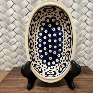 May include: An oval ceramic dish with a blue and white heart pattern. The dish has a brown rim and is decorated with small white dots. The dish is on a wooden stand.