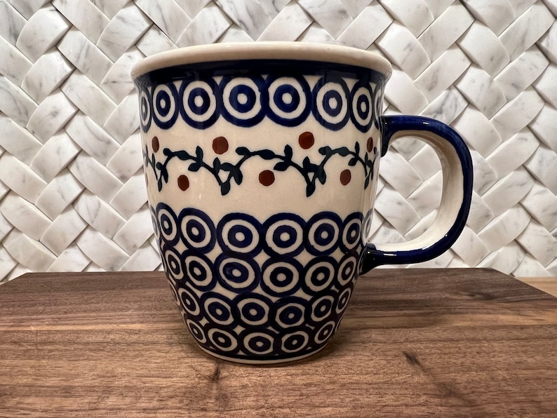 May include: A white ceramic mug with a blue and brown floral design. The mug has a blue handle and a wide, rounded base.