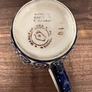 May include: A blue and white ceramic mug with a swirl pattern. The bottom of the mug has the text "HAND MADE IN ISLAND", "URA", and "MANUFACTAK" in brown lettering. The mug has a blue handle.