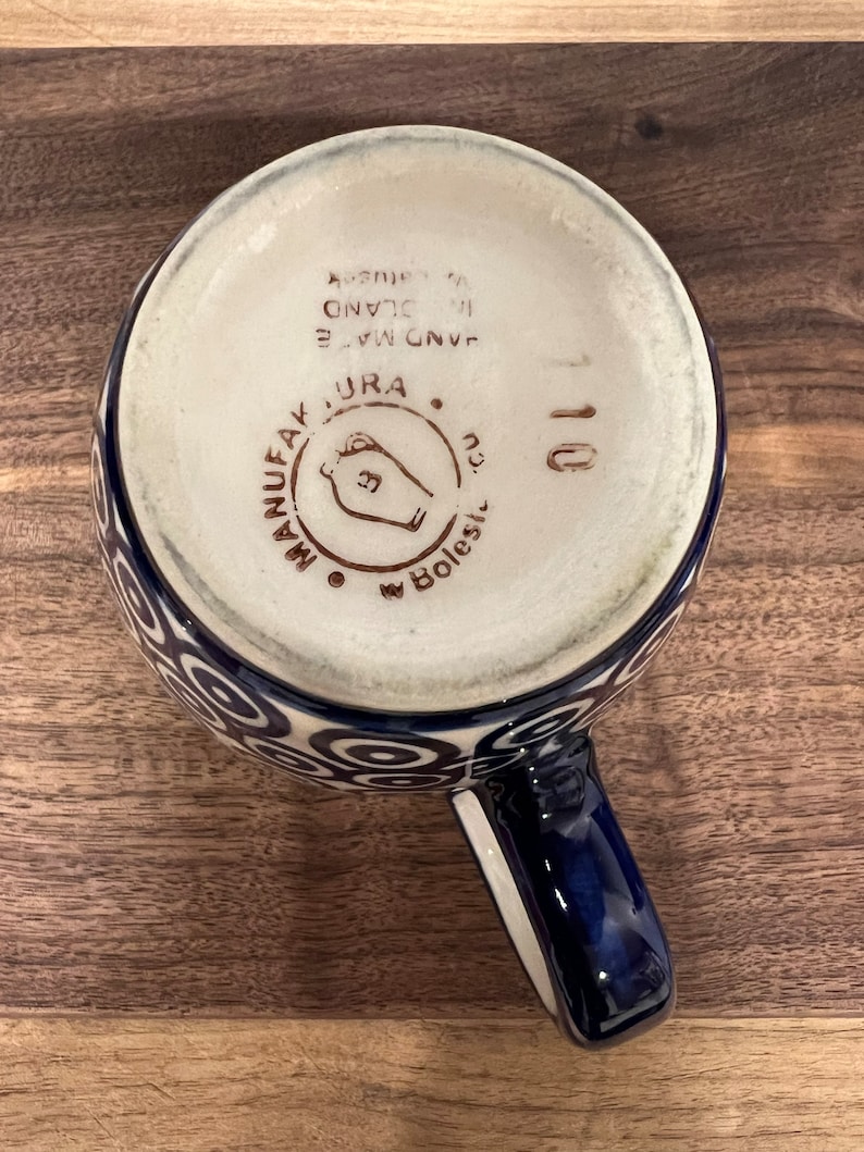 May include: A blue and white ceramic mug with a swirl pattern. The bottom of the mug has the text "HAND MADE IN ISLAND", "URA", and "MANUFACTAK" in brown lettering. The mug has a blue handle.