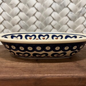 May include: A white oval ceramic bowl with a blue and brown heart pattern. The bowl has a white rim and a blue and white polka dot pattern around the bottom.