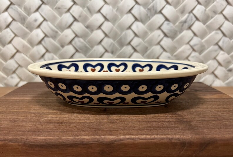 May include: A white oval ceramic bowl with a blue and brown heart pattern. The bowl has a white rim and a blue and white polka dot pattern around the bottom.