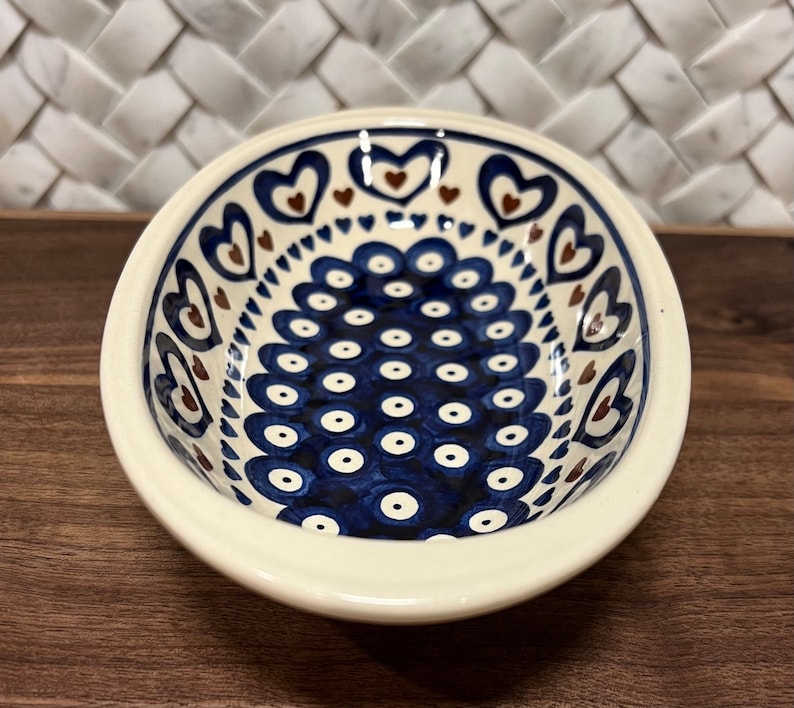 May include: A white oval ceramic bowl with a blue and brown pattern. The bowl has a repeating pattern of blue circles with white centers and a border of brown hearts.