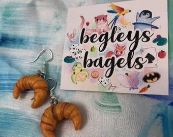 Pode incluir: Brincos em forma de croissant com ganchos prateados. Os brincos são de cor castanho dourado com riscas castanhas escuras. No fundo, um cartão com o texto "begleys bagels" e ilustrações de desenhos animados.