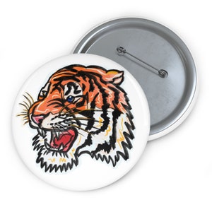 May include: A white button with a colorful illustration of a tiger's head. The tiger has orange and black stripes, a white muzzle, and a blue eye.