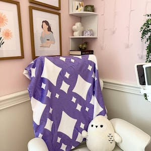 May include: A white armchair with a purple and white patterned blanket draped over it. A white owl plush toy sits on the chair. Artwork in wooden frames hangs on the pink wall behind the chair.