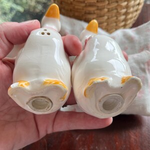 May include: A pair of white ceramic goose salt and pepper shakers with yellow beaks and feet. The shakers have pink accents and are in the shape of geese.