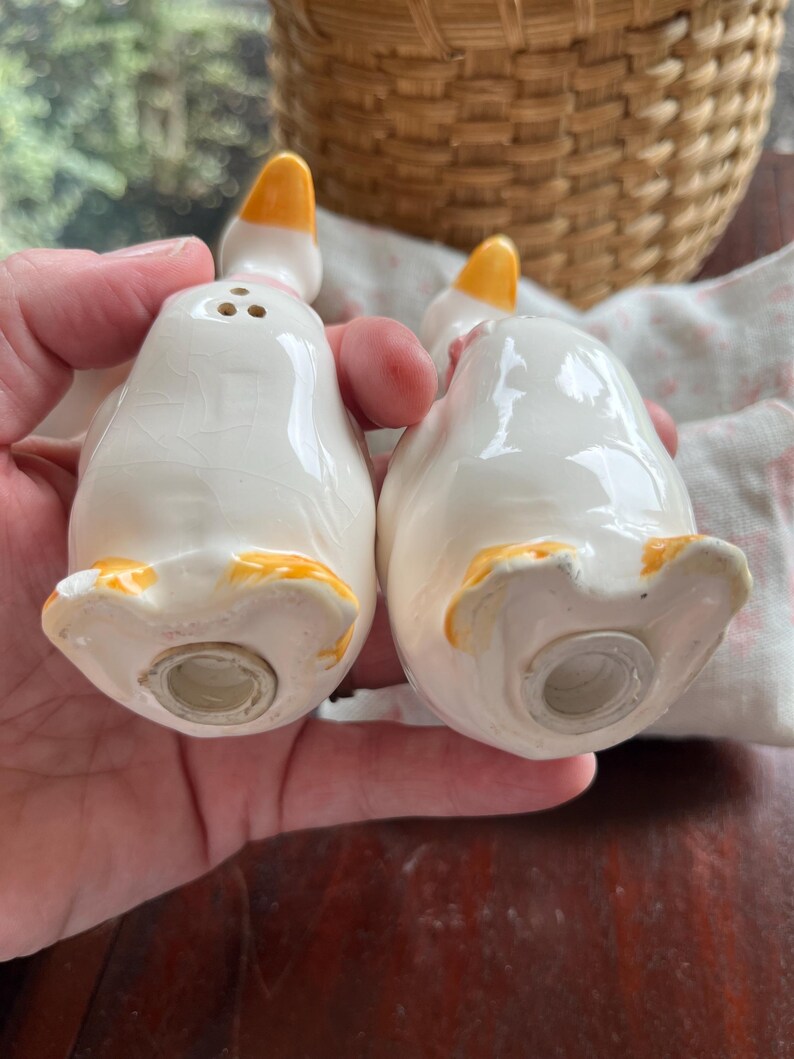 May include: A pair of white ceramic goose salt and pepper shakers with yellow beaks and feet. The shakers have pink accents and are in the shape of geese.
