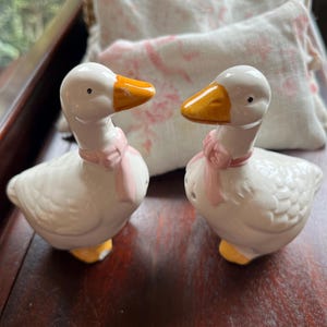 May include: A pair of white ceramic goose salt and pepper shakers with pink bows and yellow beaks.