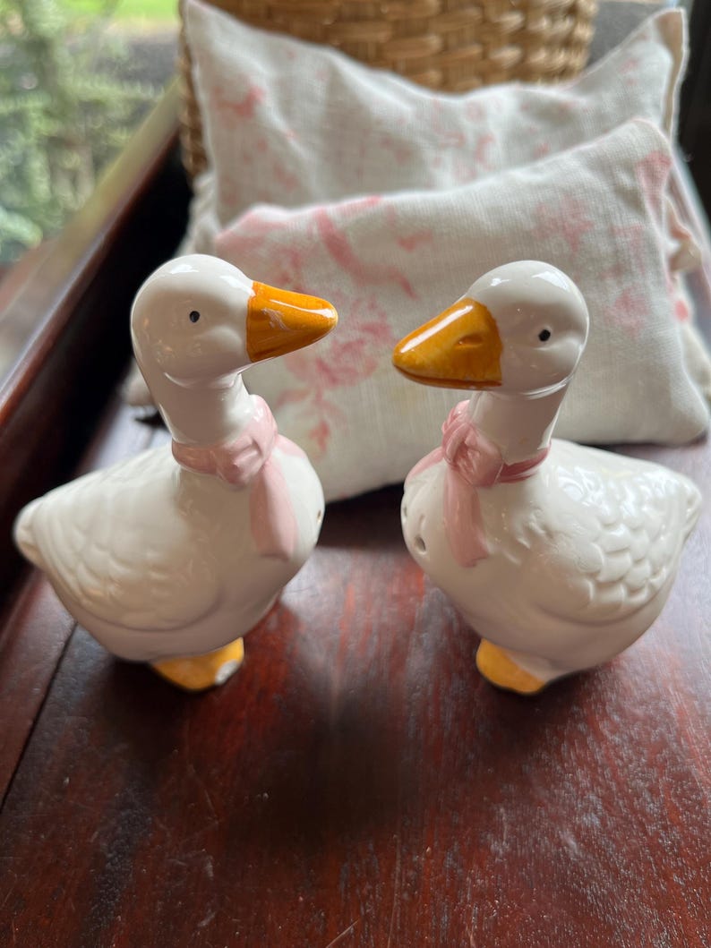 May include: A pair of white ceramic goose salt and pepper shakers with pink bows and yellow beaks.