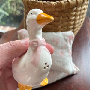 May include: White ceramic goose salt shaker with a pink bow and yellow beak and feet. The goose is standing on a table.