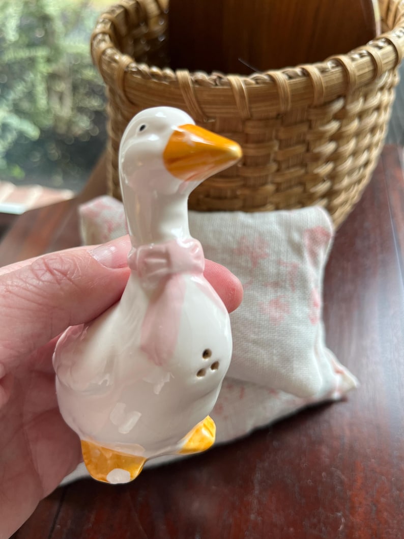 May include: White ceramic goose salt shaker with a pink bow and yellow beak and feet. The goose is standing on a table.