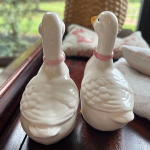 May include: Two white ceramic goose salt and pepper shakers with pink accents. The geese are facing opposite directions.