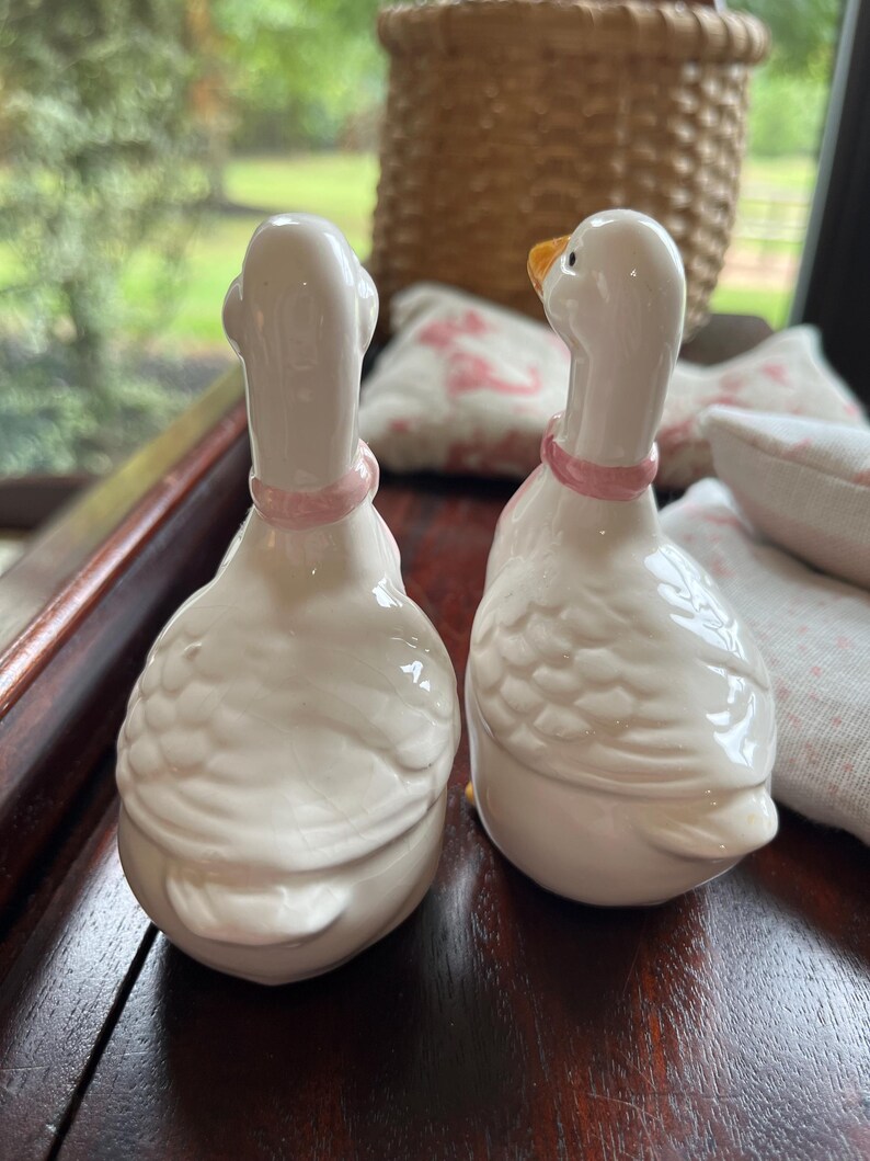 May include: Two white ceramic goose salt and pepper shakers with pink accents. The geese are facing opposite directions.