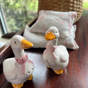 May include: Two white ceramic goose salt and pepper shakers with pink bows and yellow beaks. The shakers are on a wooden surface.