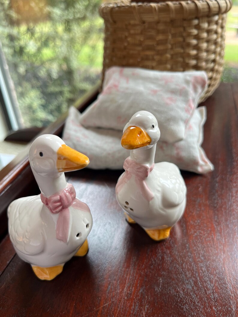 May include: Two white ceramic goose salt and pepper shakers with pink bows and yellow beaks. The shakers are on a wooden surface.