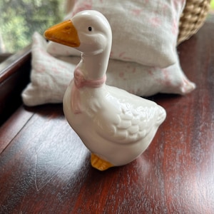May include: A white ceramic goose figurine with a pink bow around its neck. The goose has a yellow beak and feet and is standing on a wooden surface.