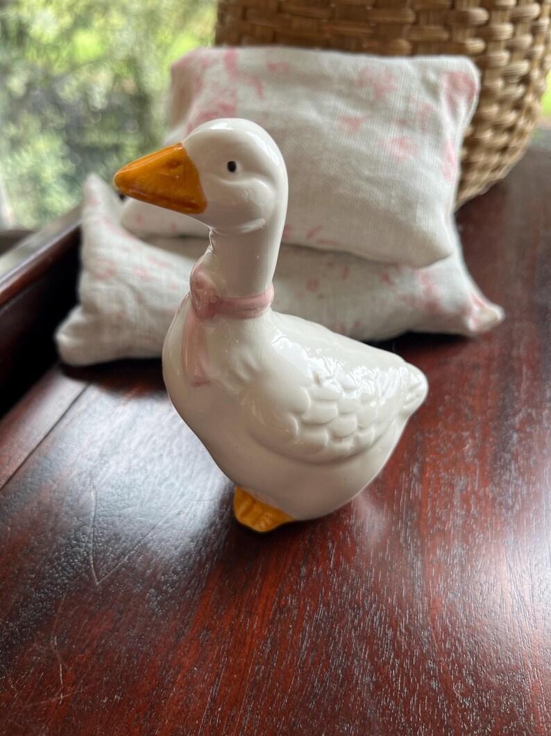 May include: A white ceramic goose figurine with a pink bow around its neck. The goose has a yellow beak and feet and is standing on a wooden surface.