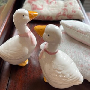 May include: Two white ceramic goose figurines with pink ribbons around their necks. The geese have yellow beaks and feet. The figurines are on a wooden surface.