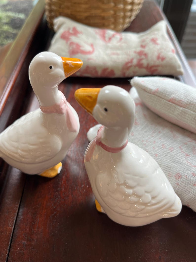May include: Two white ceramic goose figurines with pink ribbons around their necks. The geese have yellow beaks and feet. The figurines are on a wooden surface.