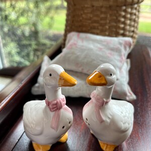 May include: A pair of white ceramic goose salt and pepper shakers with pink bows. The shakers have yellow beaks and feet.